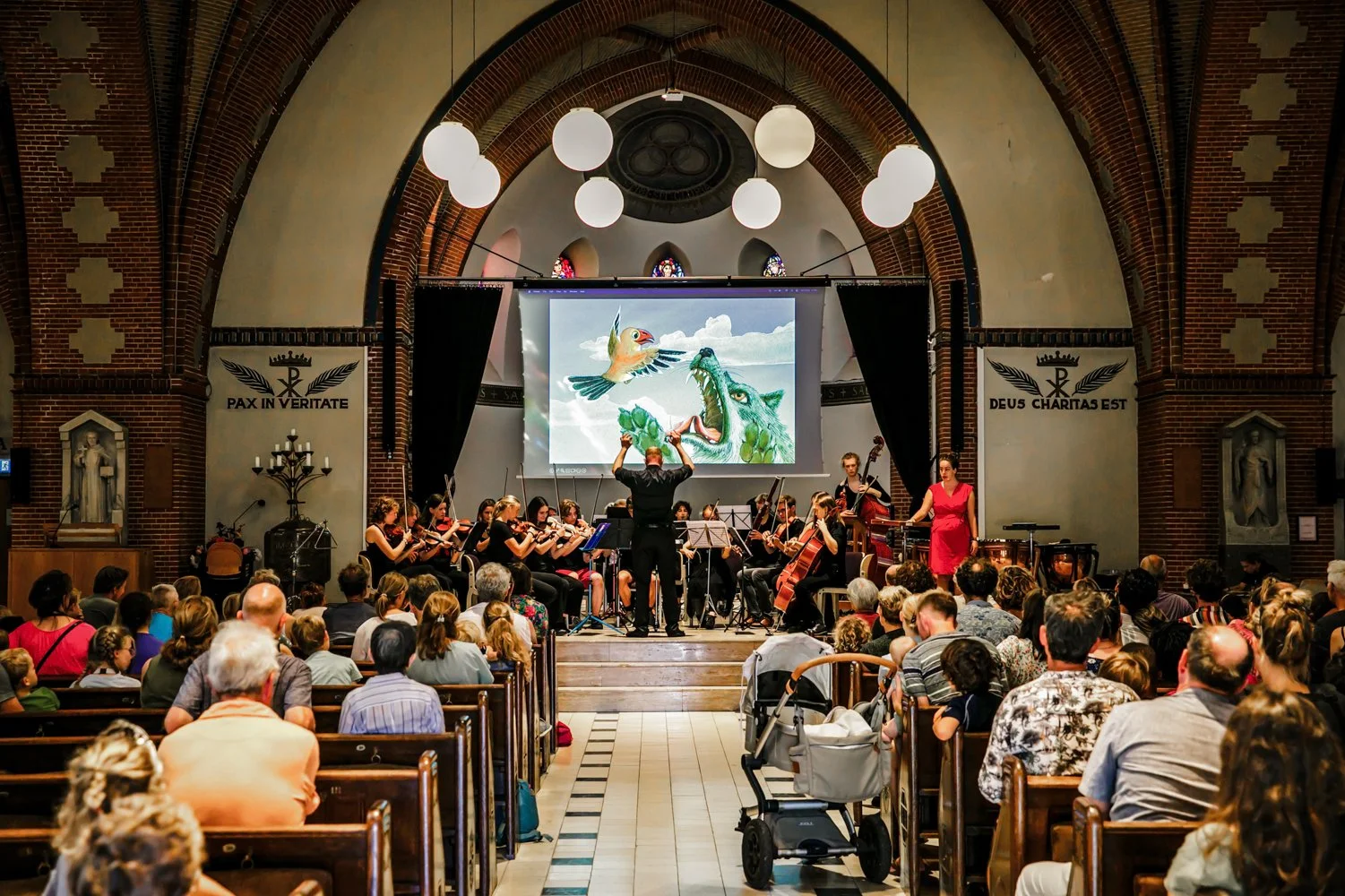 An orchestra is performing in a church with a conductor, two women, and musicians with various instruments, facing an audience seated in benches, with a large screen displaying colorful animated images of a bird and a dragon.