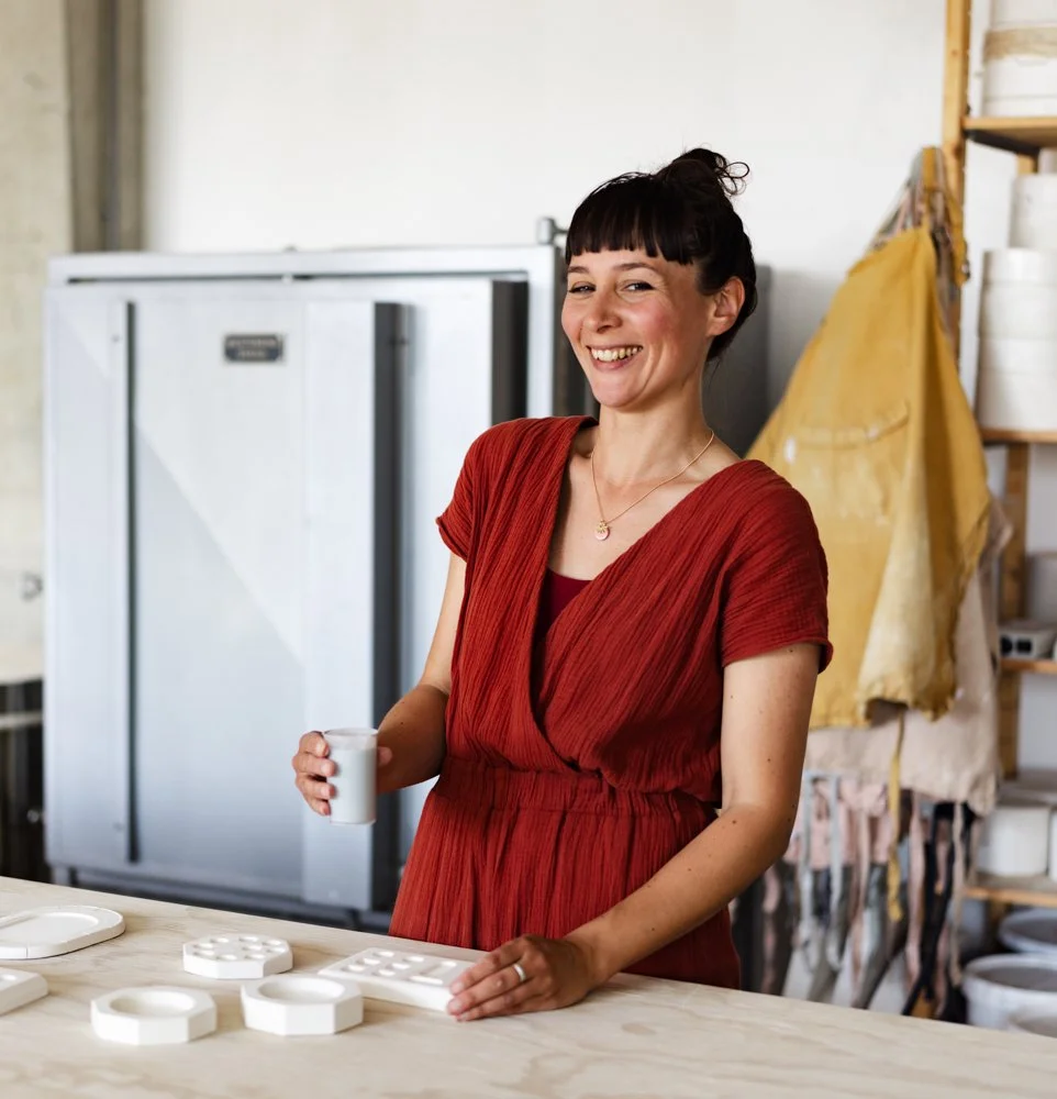 Vrouw in een rood jurk die in een atelier of keuken glimlachend een glas melk vasthoudt, omringd door witte keramische objecten.