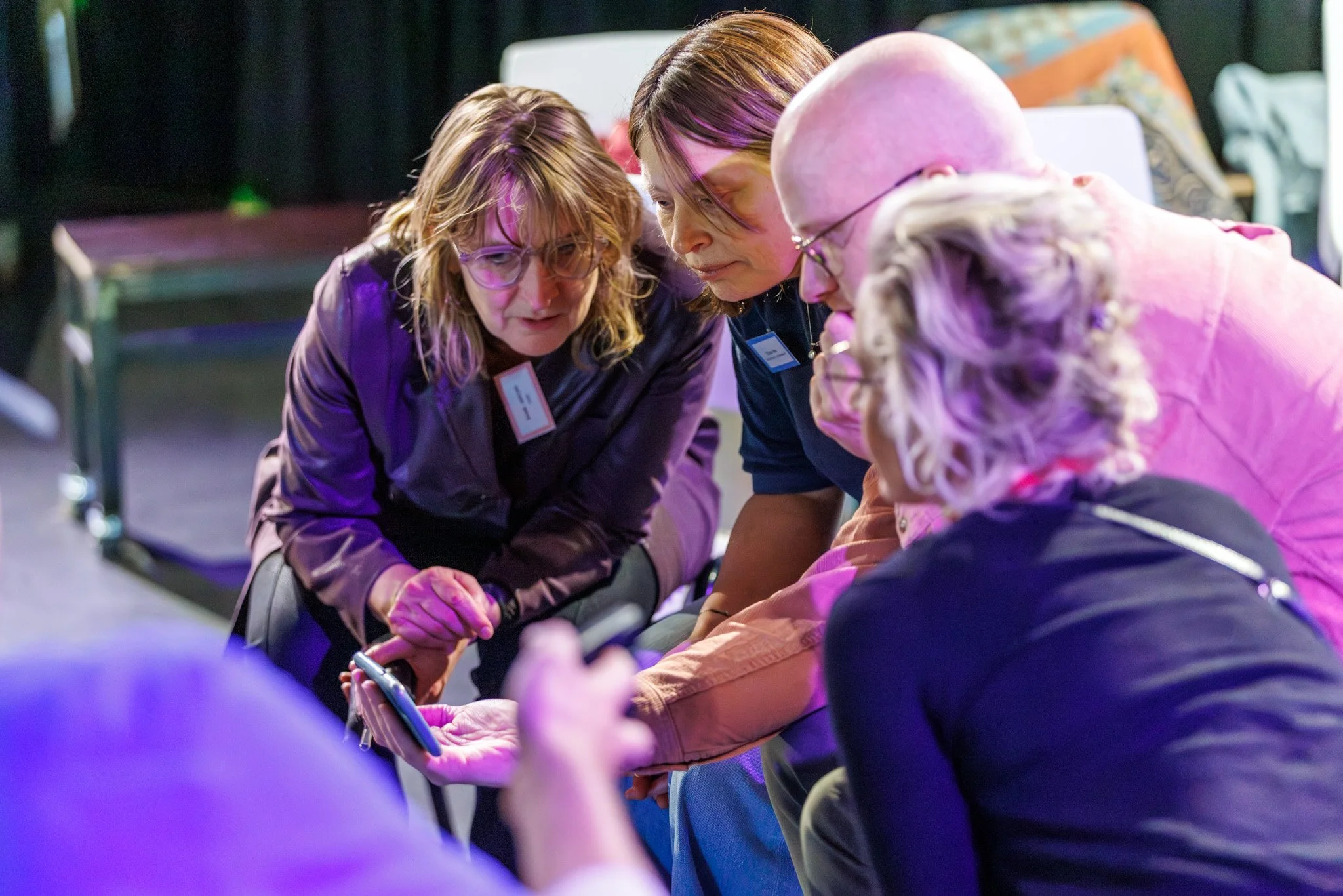 Four women with short and shoulder-length hair, wearing glasses and name tags, are closely gathered and leaning in to look at a smartphone, in a dimly lit indoor setting with purple lighting.