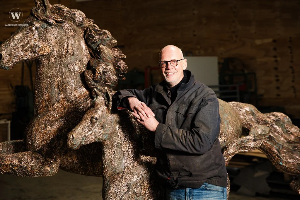 Man met bril, zwart jack, blauwe jeans, staat naast een grote bronzen paardensculptuur in een houten ruimte.