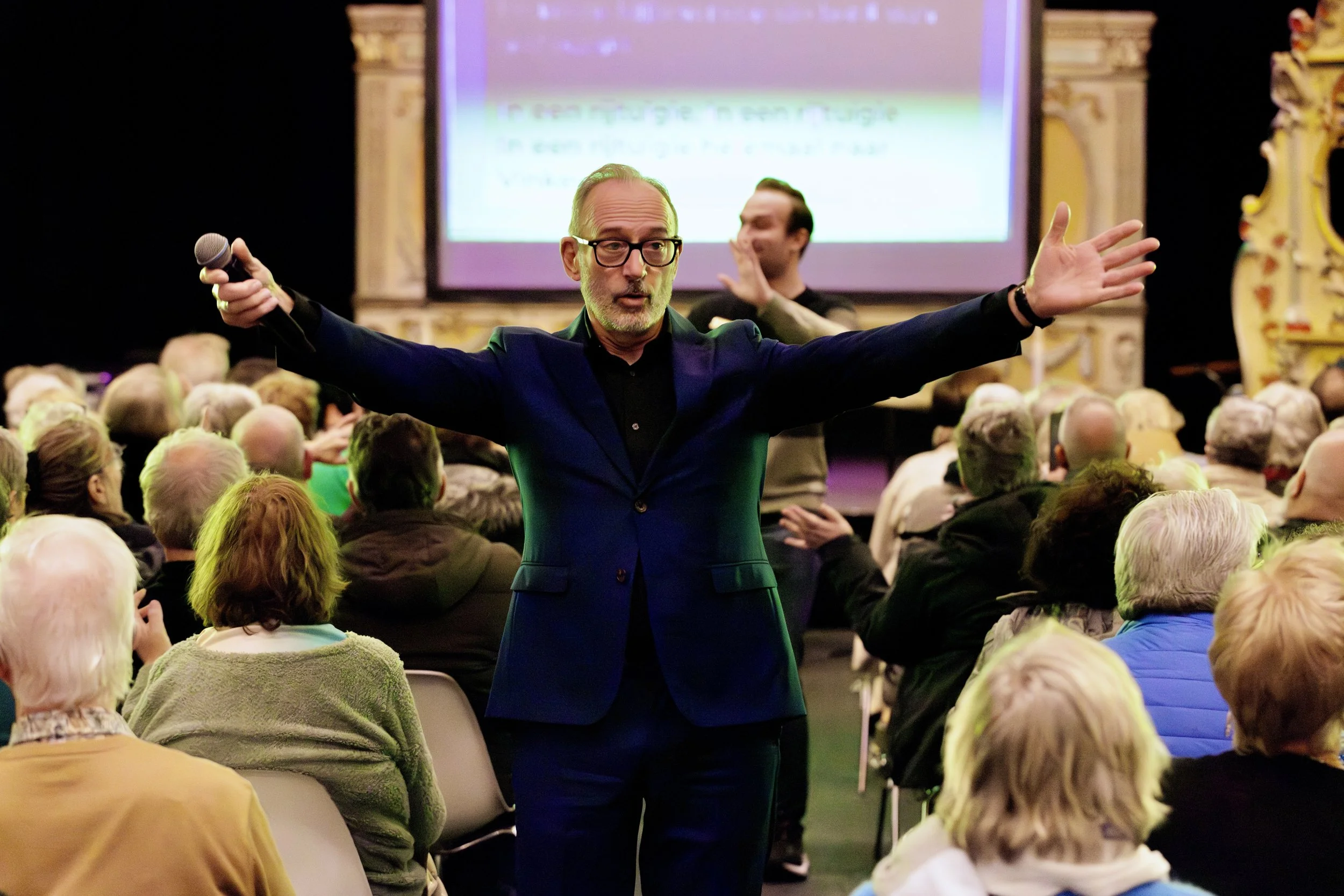 A man with glasses in a blue suit speaking to an audience with his arms wide open in a conference room, with a large screen and decorative frame behind him, and people seated facing him.