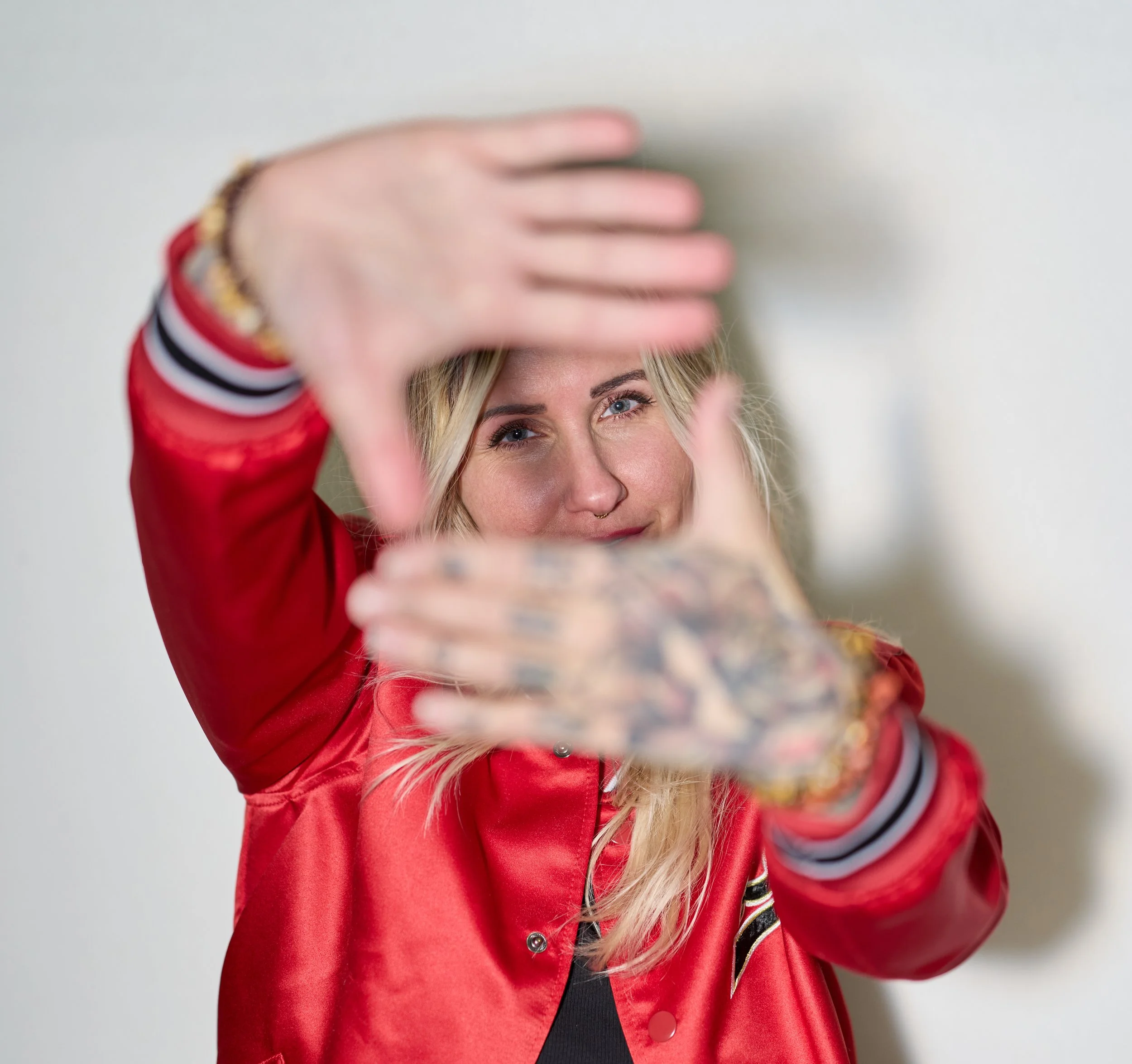 Close-up of a woman with blonde hair and blue eyes, wearing a red varsity jacket with black and white striped cuffs, taking a selfie with a blurred phone and background.