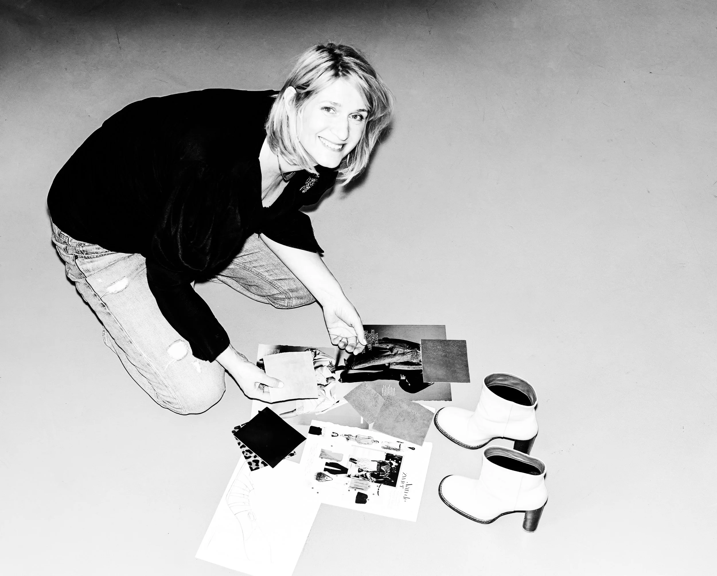 A woman crouching down, holding a photograph, with various papers, magazines, and a pair of boots on the floor, against a plain background.