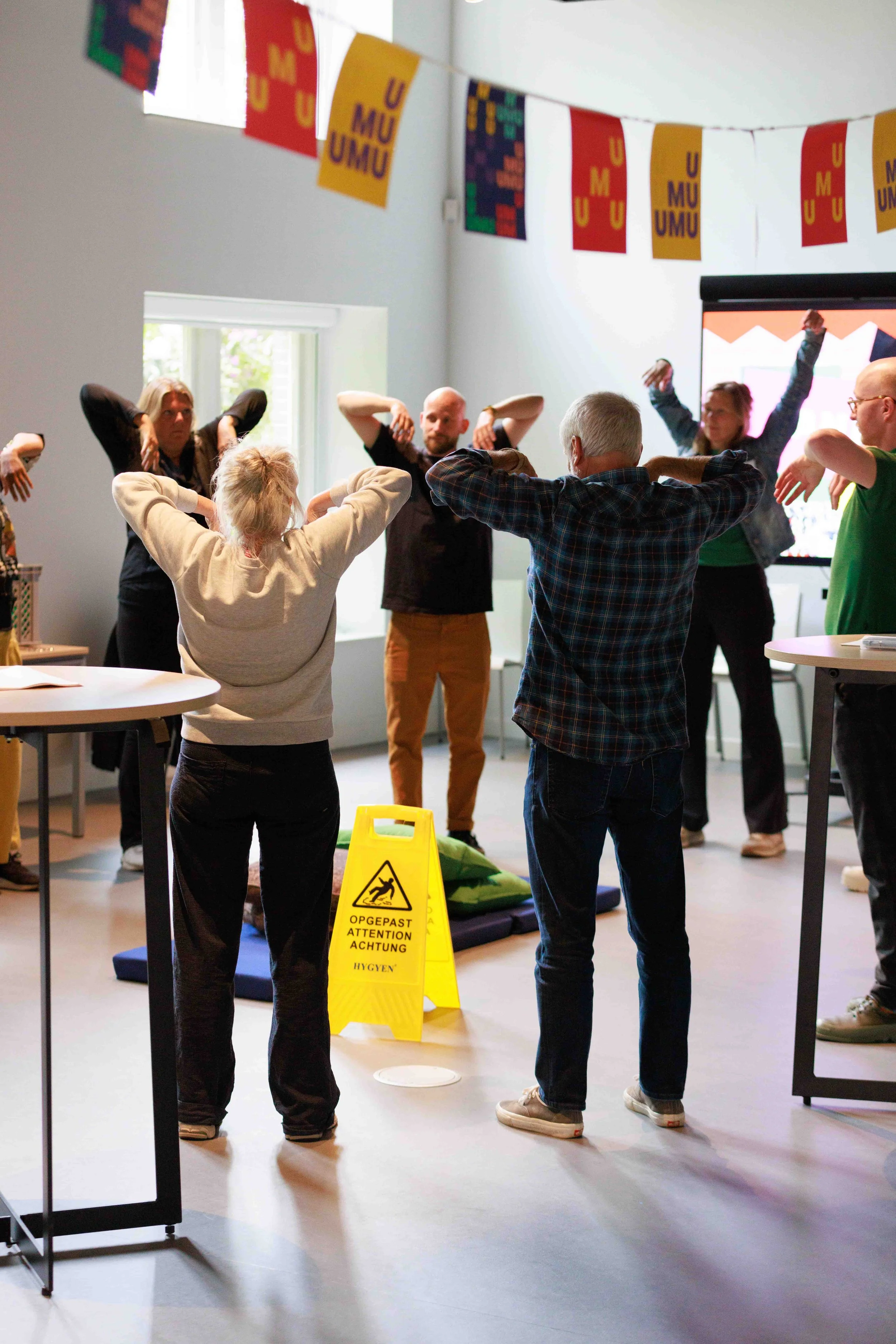 People participating in a group exercise class indoors, with some stretching their arms and a new person at the front demonstrating. A yellow caution sign on the floor indicates wet floor, and decorations with 
