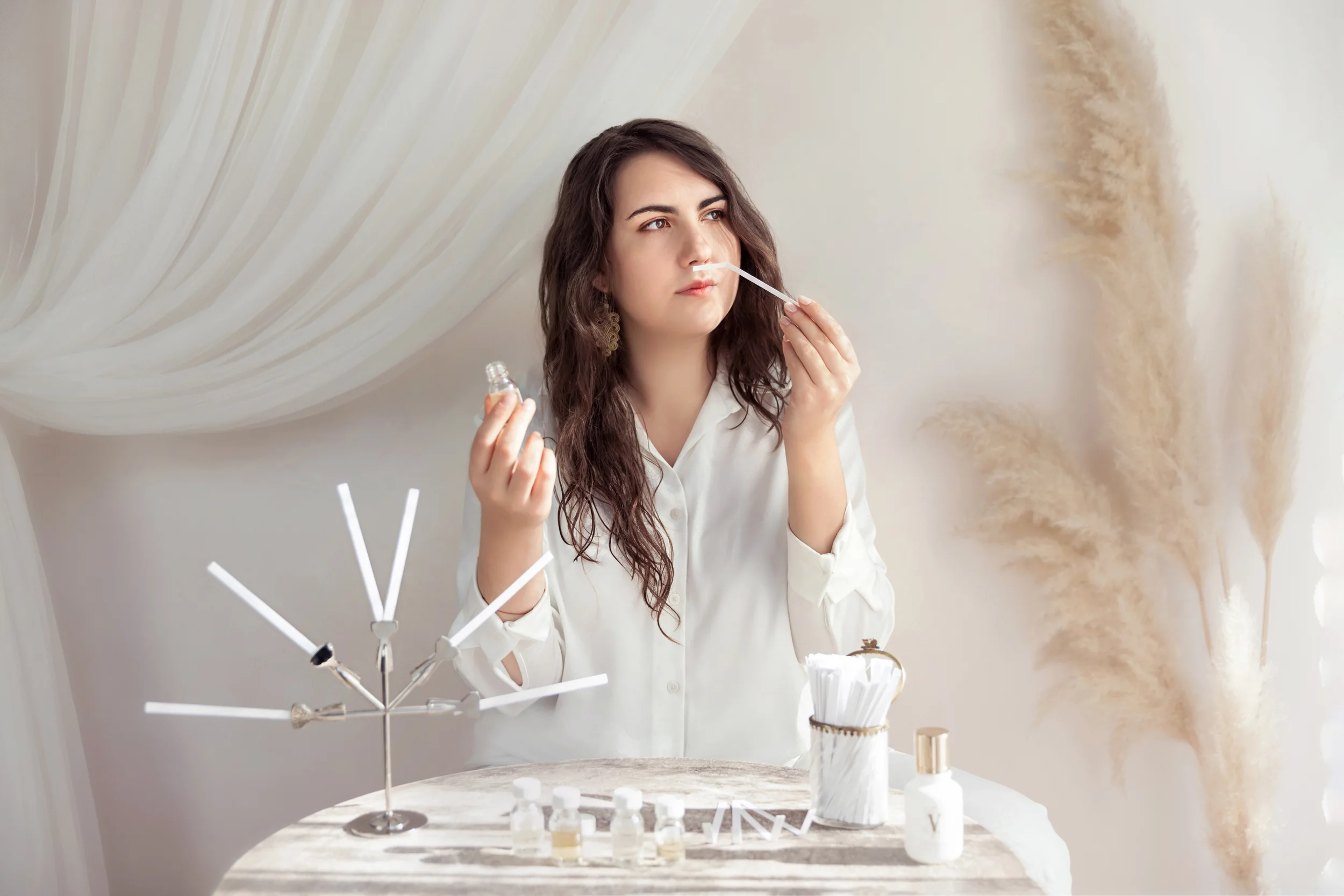 A young woman with wavy brown hair and earrings applies nasal spray at a marble table with various bottles and containers, in a room with a neutral textured wall.