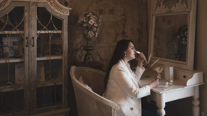 A woman in a white blazer is sitting at a white vanity table, looking out of a window, holding a makeup brush near her face, with a mirror, small bottles, and a lamp on the table. The room has antique-style furniture and floral decor.