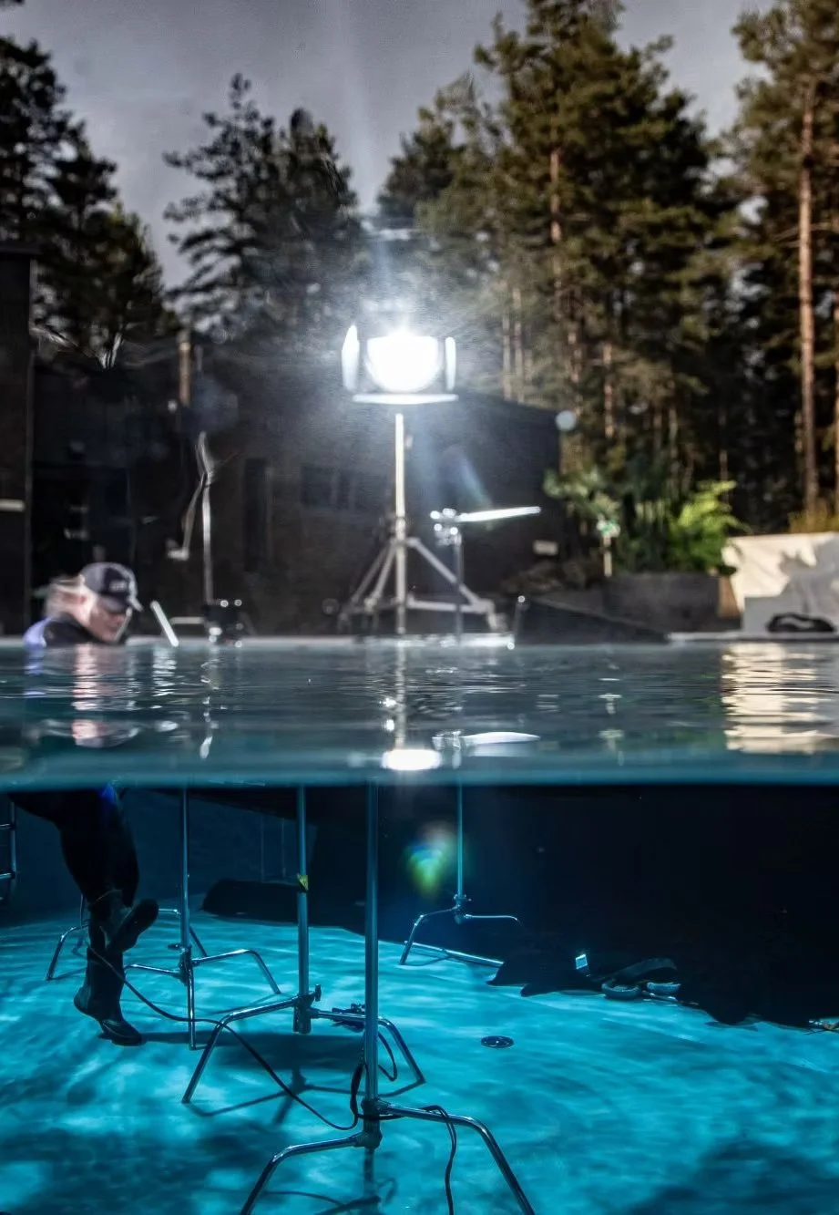 An outdoor filming set under a large bright light with trees in the background. The shot is taken partly underwater, showing underwater equipment and a person working below the surface.