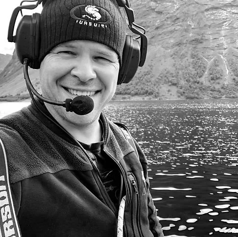 Person smiling with headphones and a microphone, outdoors by a body of water with mountains in the background.