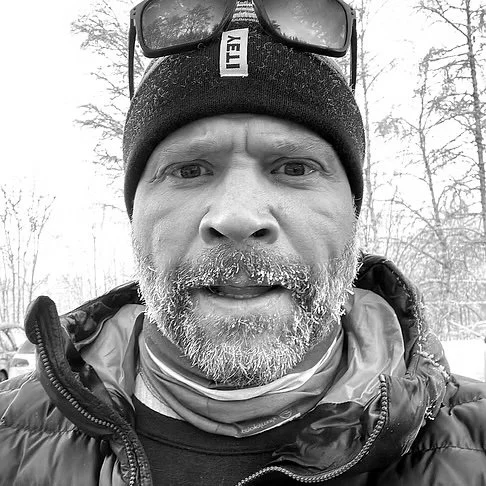 A man with a gray beard wearing a black beanie, sunglasses on his forehead, a jacket, and a neck gaiter, standing outdoors in a snowy winter landscape.