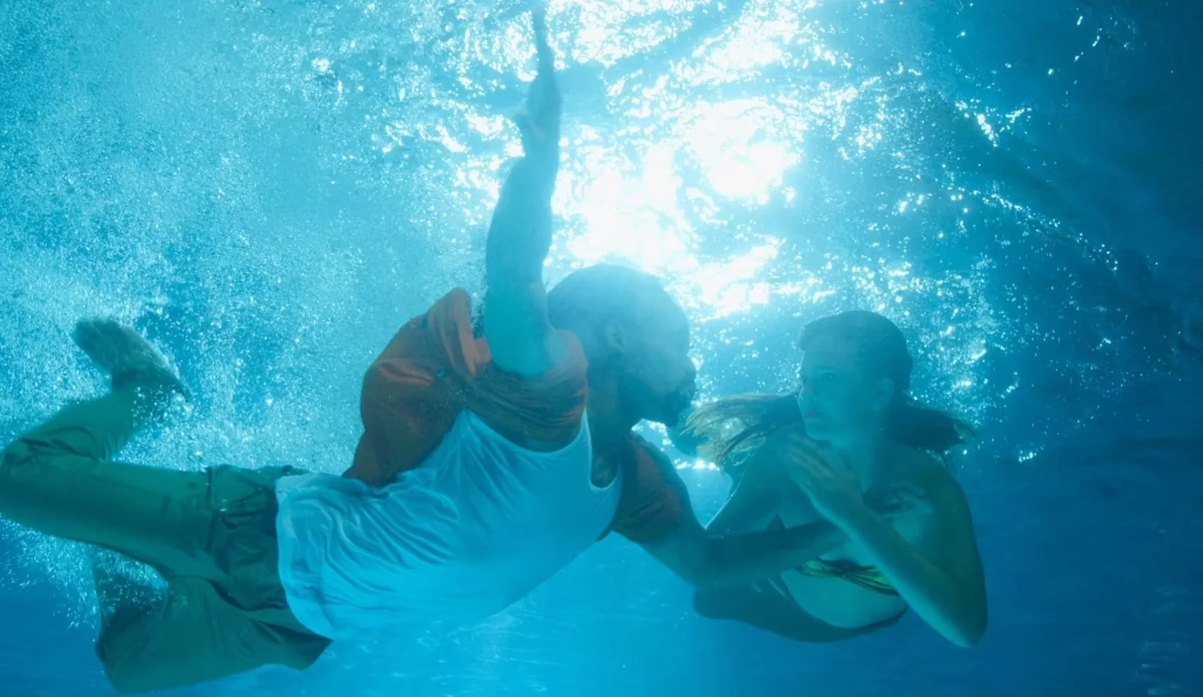 Two people are underwater, engaged in a struggle or fight. One person appears to be grabbing the other, with bubbles surrounding them and sunlight piercing through the water surface above.
