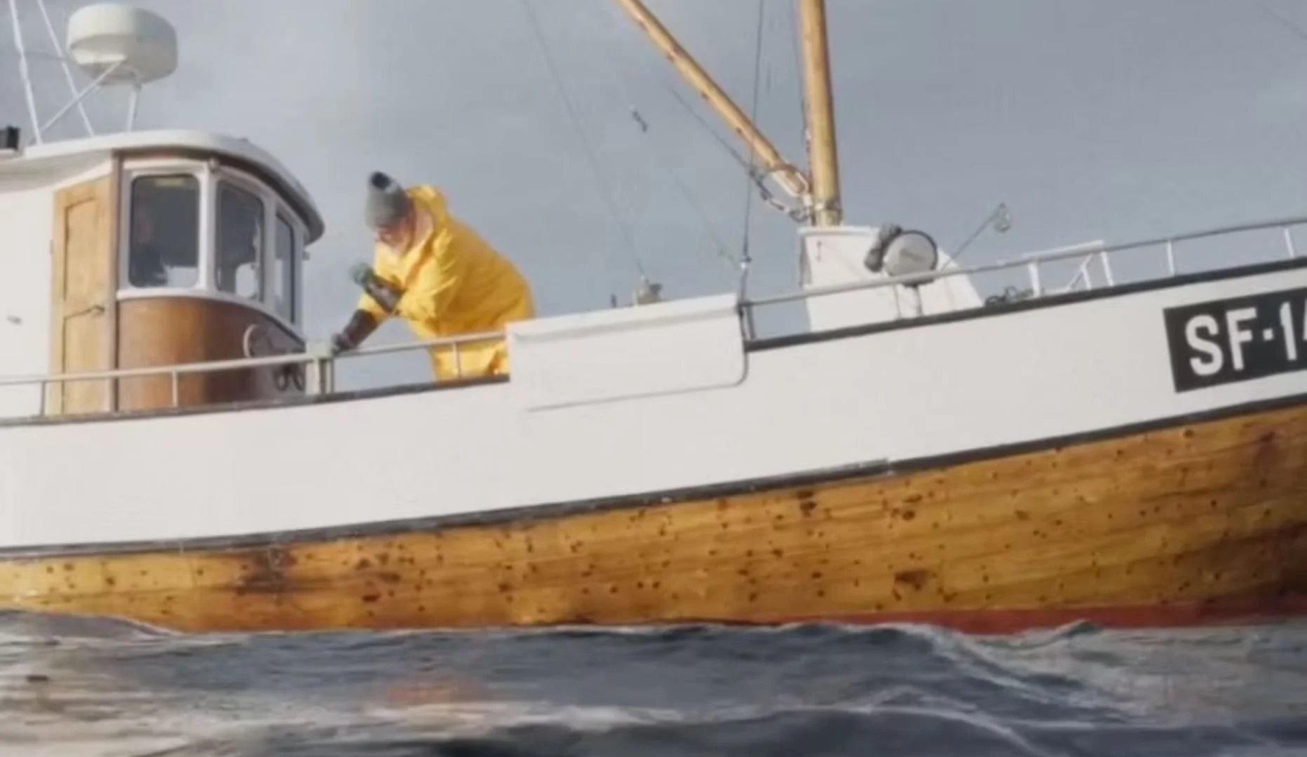 A person in a yellow raincoat is standing on a boat with a wooden hull and white upper structure, seen from a lower angle with water in the foreground.