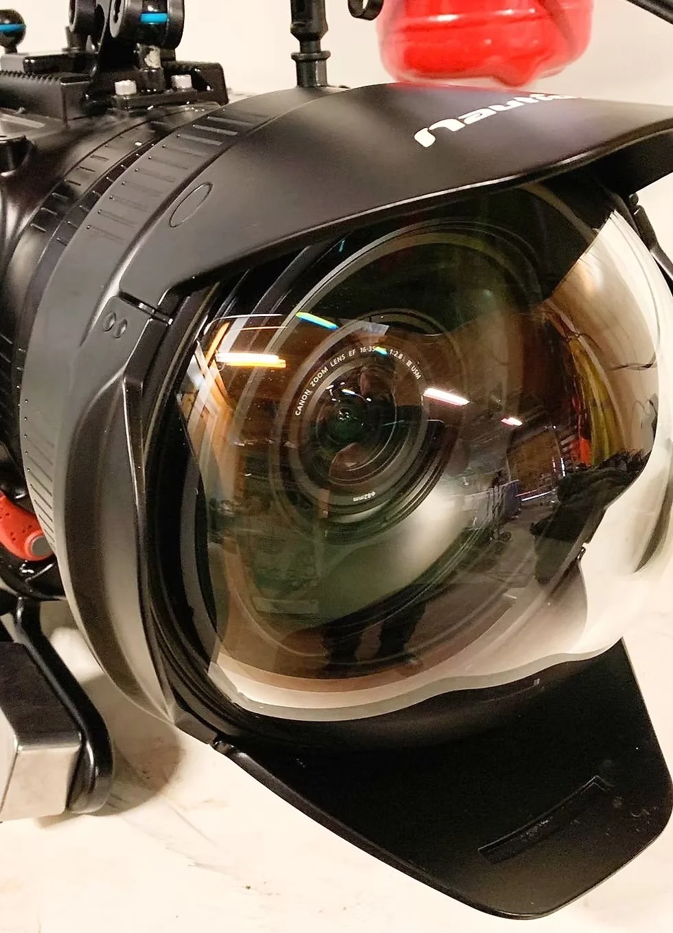 Close-up of a professional camera lens mounted on a camera body, with a red object and some mechanical parts visible in the background.