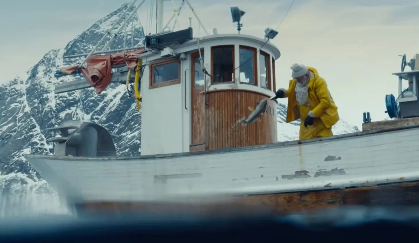 A person in yellow rain gear and a white hat is holding a fish on a boat in an icy, snowy landscape with mountains in the background.