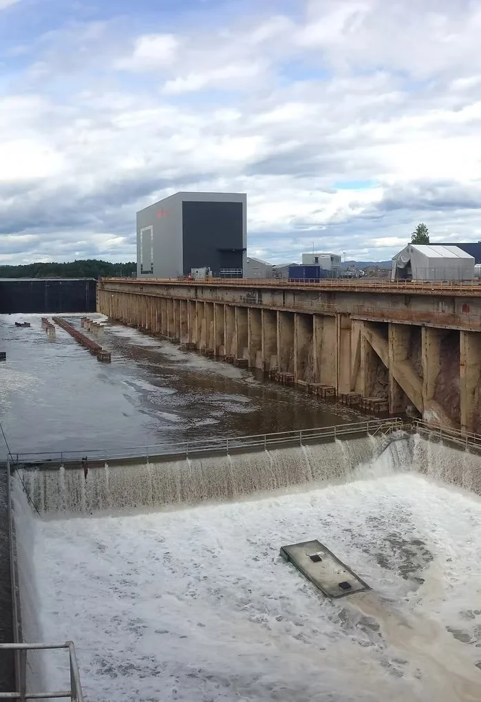 Hydroelectric dam with water flowing over the spillway, and industrial buildings in the background under a cloudy sky.