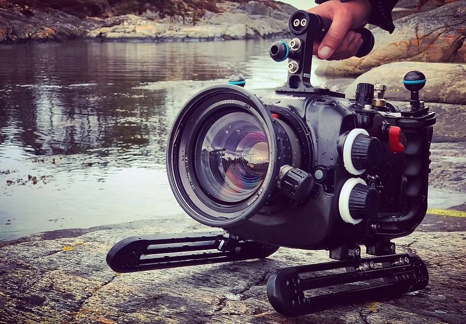 A professional underwater camera with a large lens, mounted on a track-style underwater housing, positioned on rocks by a river or lake shoreline with rocks and water in the background.