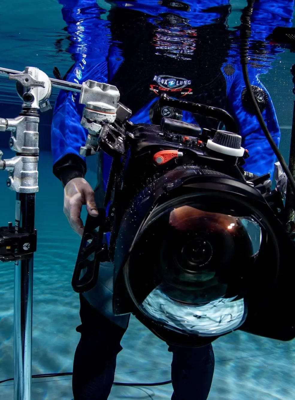 A diver underwater holding a professional underwater camera rig with a large lens, wearing a blue wetsuit with the logo 'Aqua Lung'.