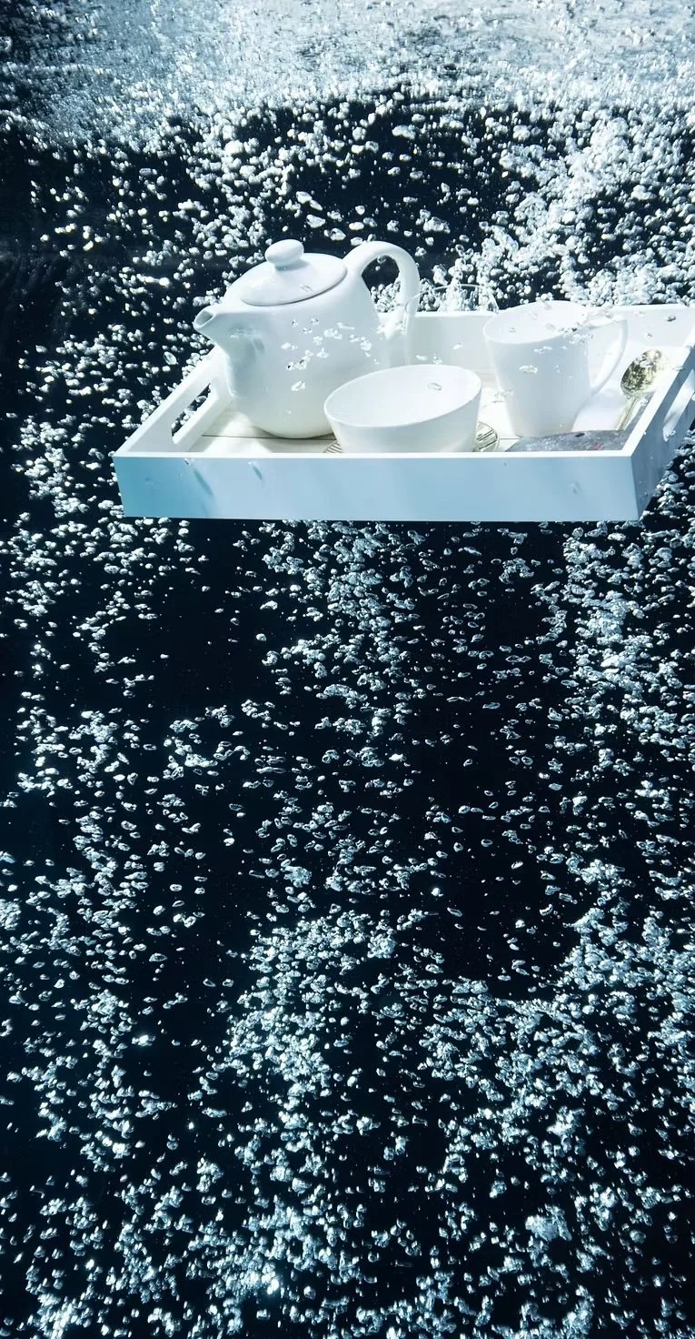 A white tray with a teapot, cup, plate, and spoon submerged in dark water with many air bubbles.