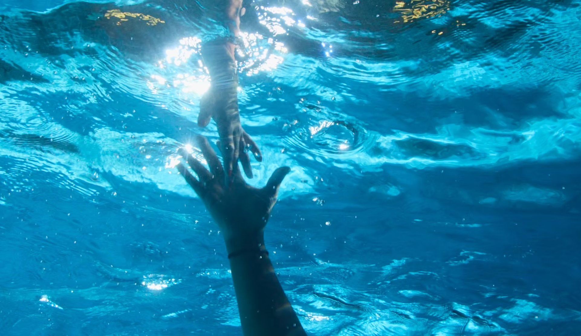 A hand reaching out of a swimming pool with sunlight reflecting on the water surface.