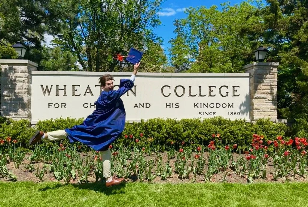 A graduate in a blue cap and gown jumping happily in front of Wheaton College sign on a sunny day with trees and tulips in the background.