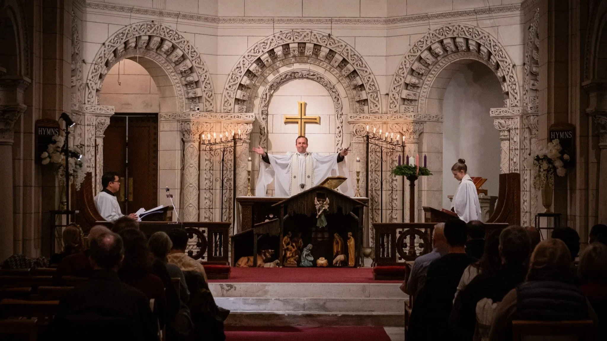 A church service with a priest standing at the altar, arms outstretched, with two altar servers on either side, in front of a congregation. The church has ornate architecture with a cross and candelabras visible.