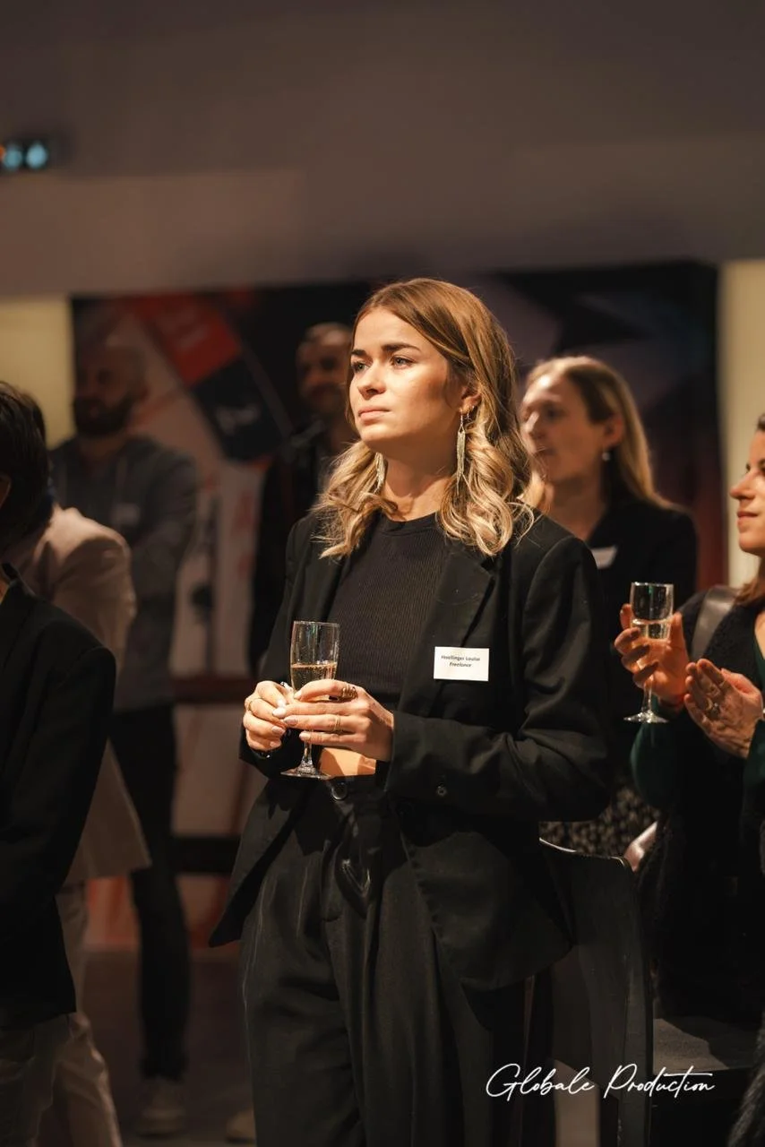 Femme tenant une coupe de champagne lors d'une réception professionnelle, portant une veste noire et une tenue élégante, dans une ambiance de networking ou de célébration.