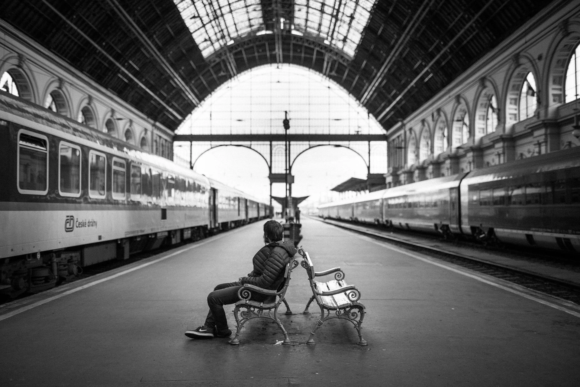 Black and white photograph of a person sitting alone between trains in a large station, evoking distance, memory and separate paths