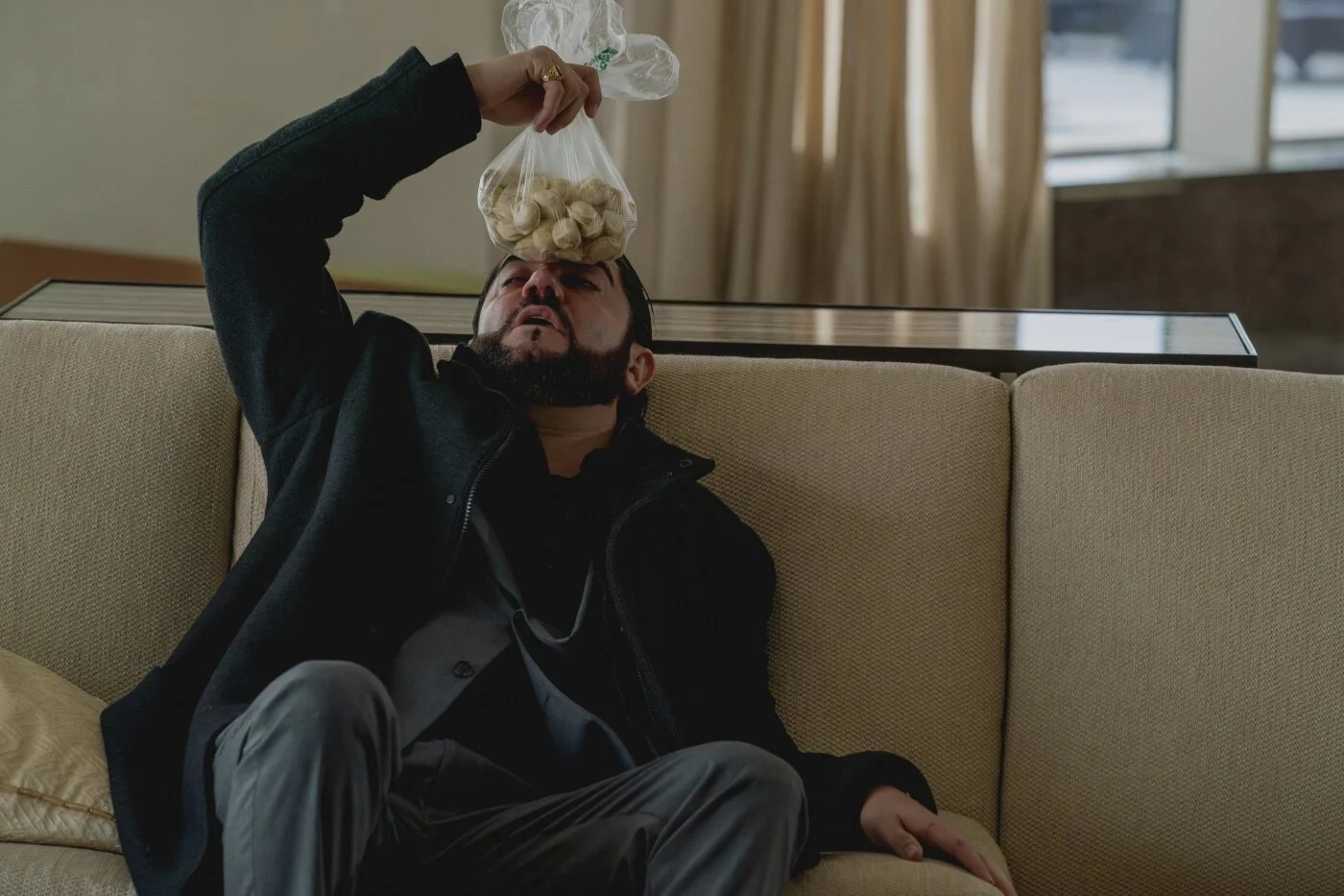 Igor’s injured cousin sitting on a sofa, holding a bag of ice to his forehead after the apartment fight in Anora.