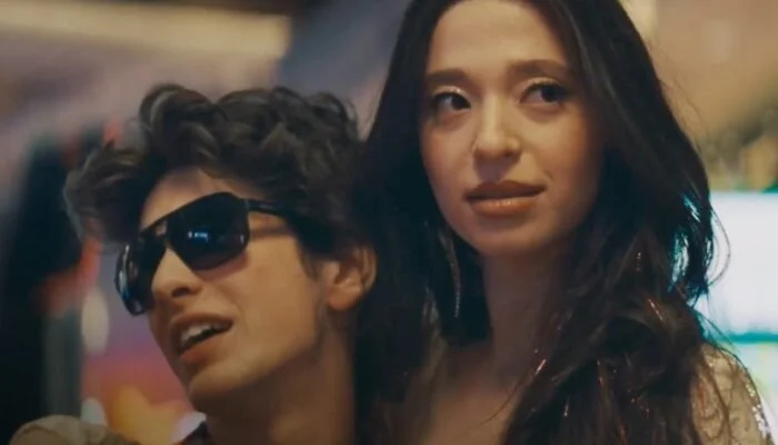 Ani and Ivan in a casino, tightly framed; she looks distant while he leans into her wearing sunglasses.