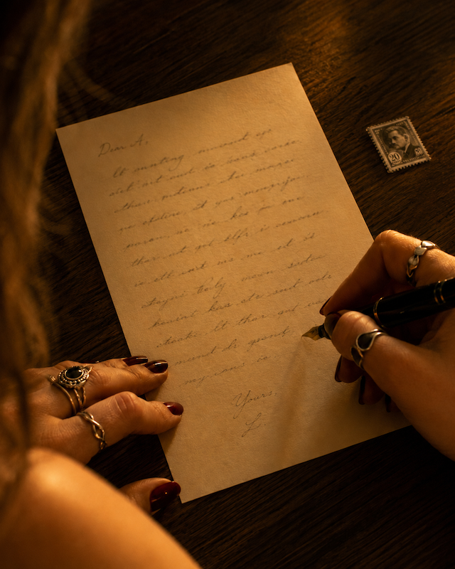 Close-up of a woman’s hands with dark cherry nails and silver rings writing a letter with a fountain pen in warm amber light, with a postage stamp nearby.
