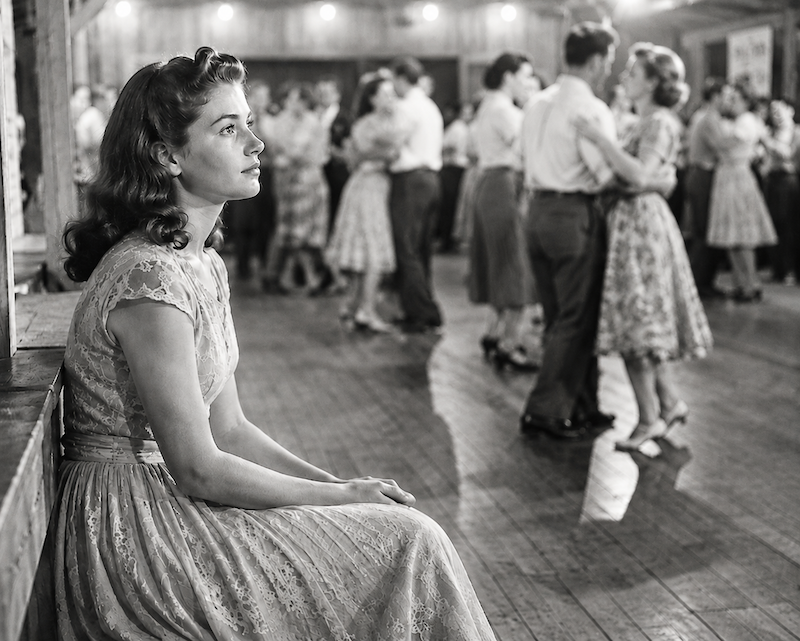 Young woman sitting alone at the edge of a 1950s dance hall while couples dance together behind her.