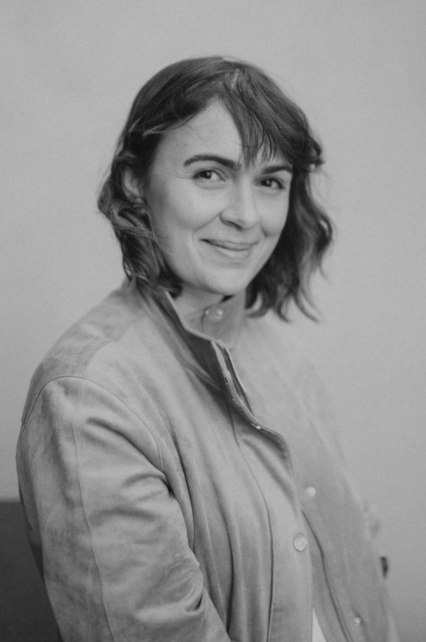 Black and white portrait of a woman with wavy hair, smiling softly, wearing a zip-up jacket, standing against a plain background.