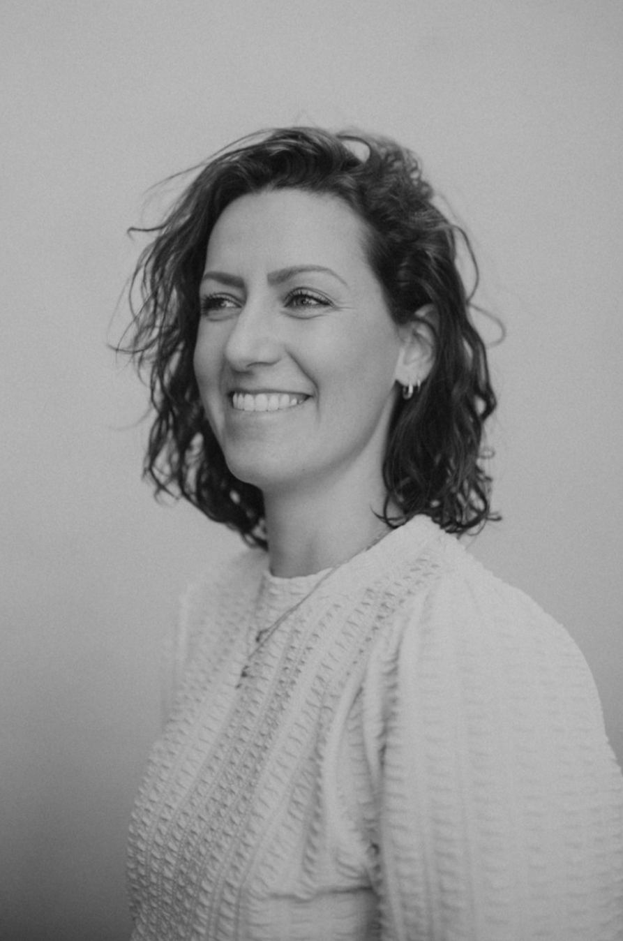 Black and white portrait of a smiling woman with curly hair, wearing earrings and a textured sweater, looking to the side.