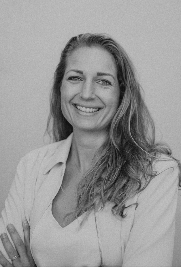 A smiling woman with long wavy hair, wearing a light-colored top and blazer, standing against a plain background.