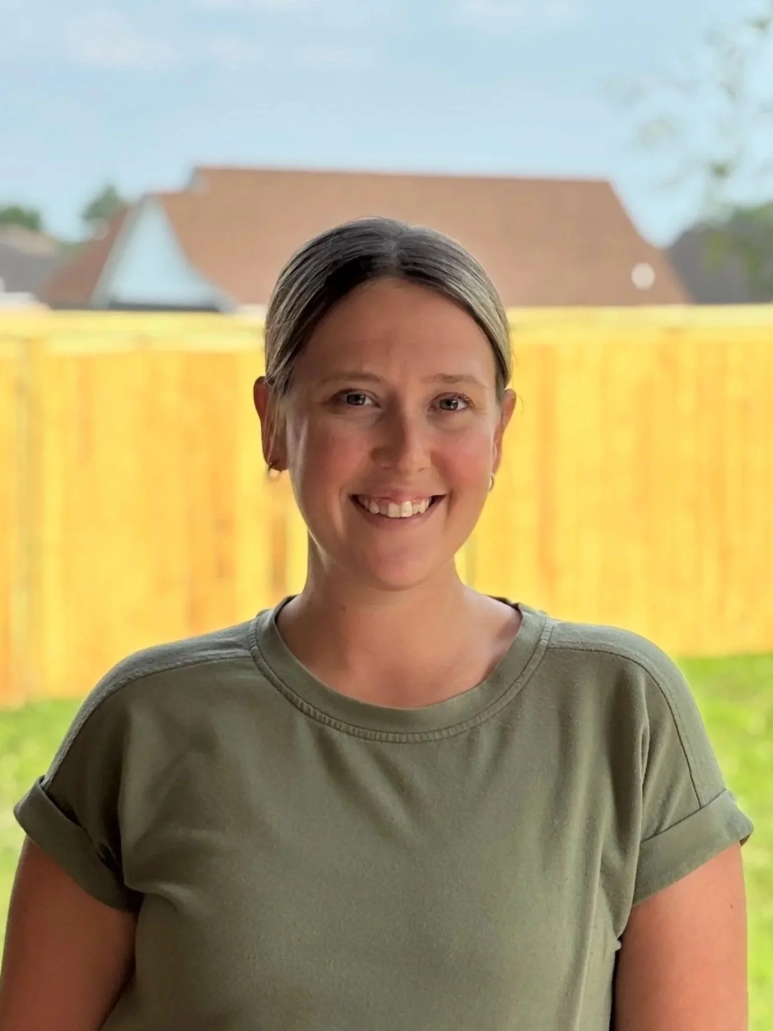 A woman with short hair and a big smile stands outdoors in front of a yellow gate, with a house and blue sky in the background.