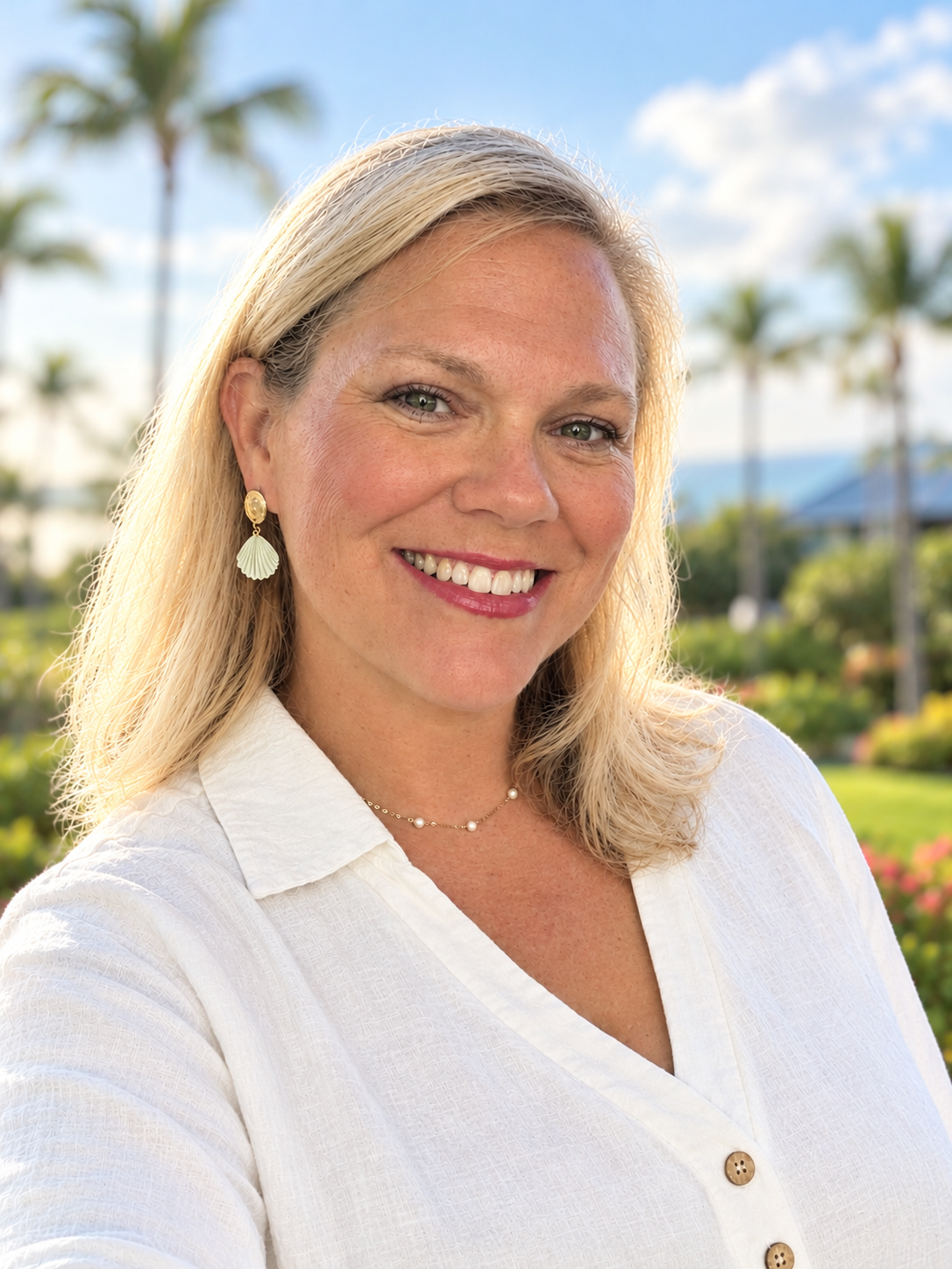 A smiling woman with blonde hair wearing a white blouse, with a background of palm trees and a blue sky.