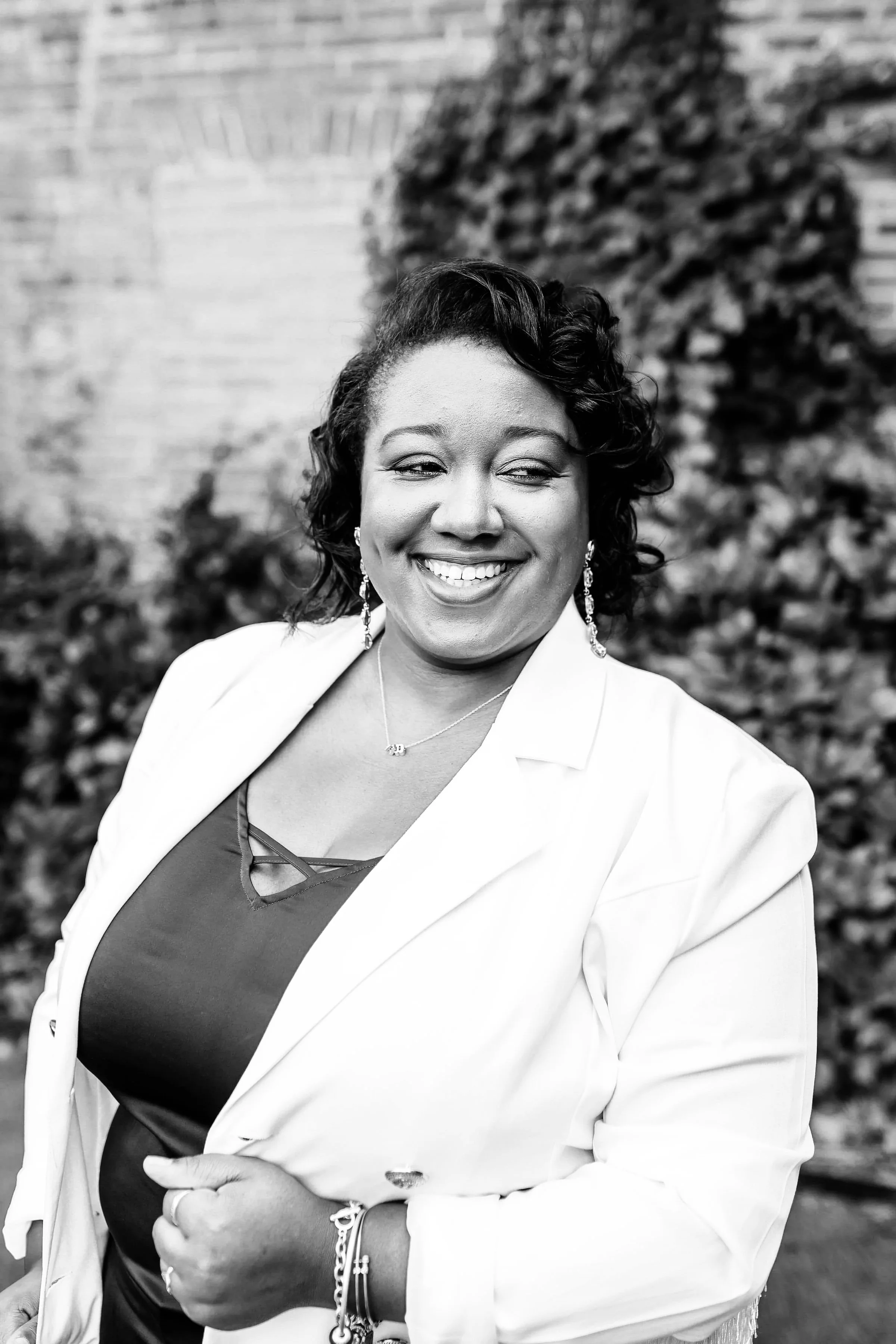 Black and white photo of a smiling woman with short curly hair, wearing earrings, a necklace, a dark top with a crisscross neckline, and a white blazer, standing outdoors in front of a brick wall and foliage.