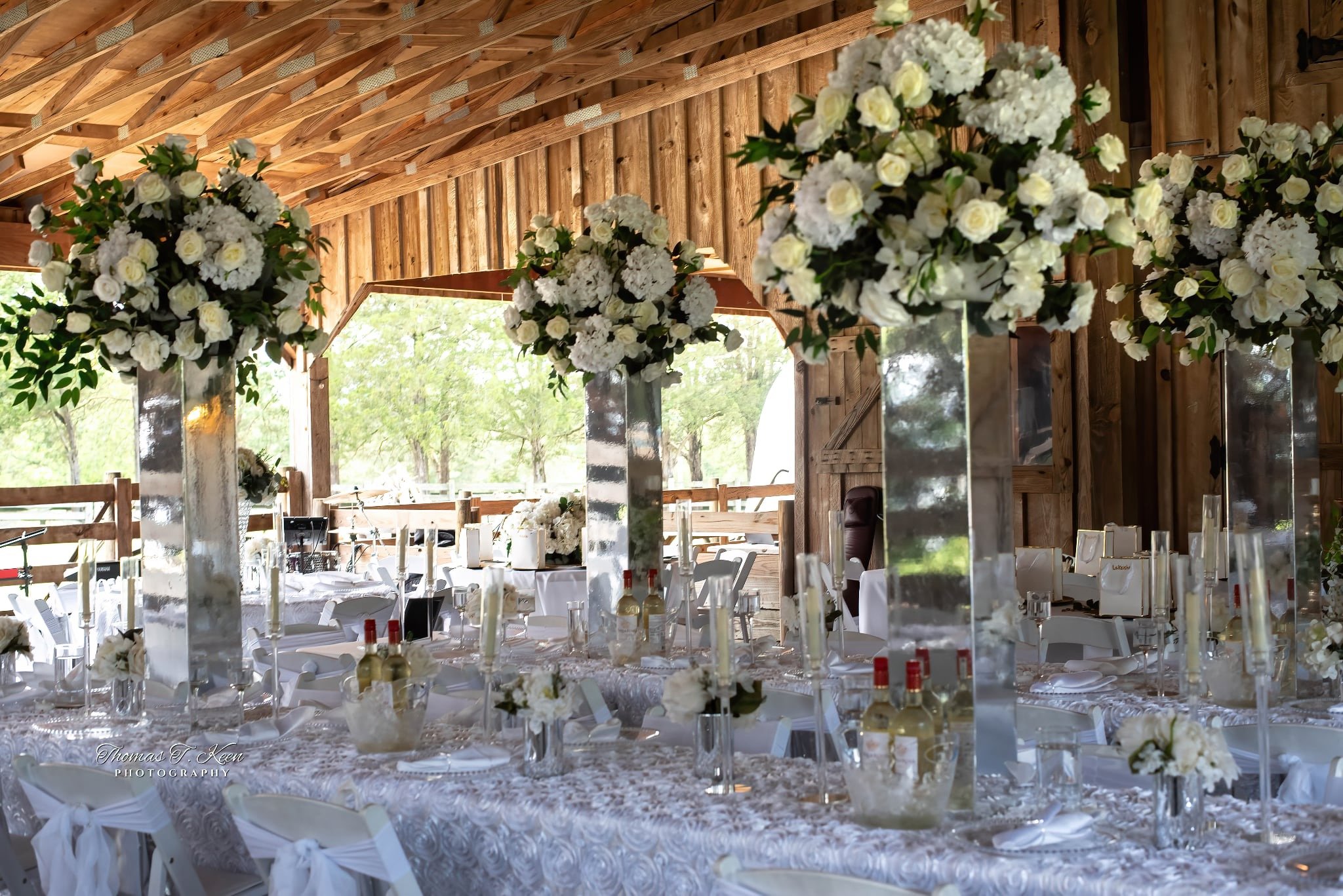 Elegant wedding reception in a rustic barn with floral centerpieces, white tablecloths, and wooden walls, overlooking an outdoor green landscape.