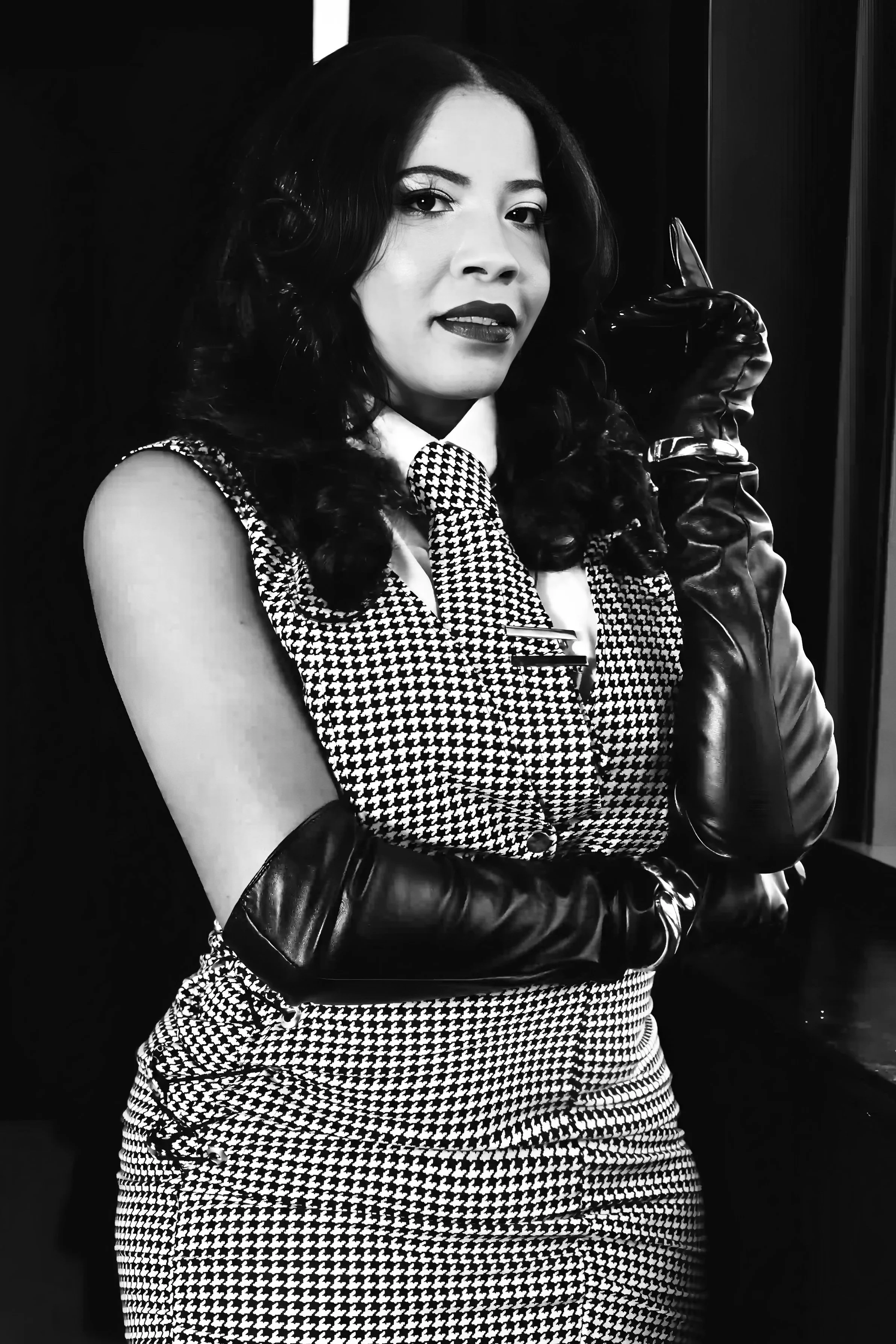 A woman with dark curly hair, wearing a houndstooth dress and matching tie, poses with her arms crossed wearing long black leather gloves, in black and white photography.