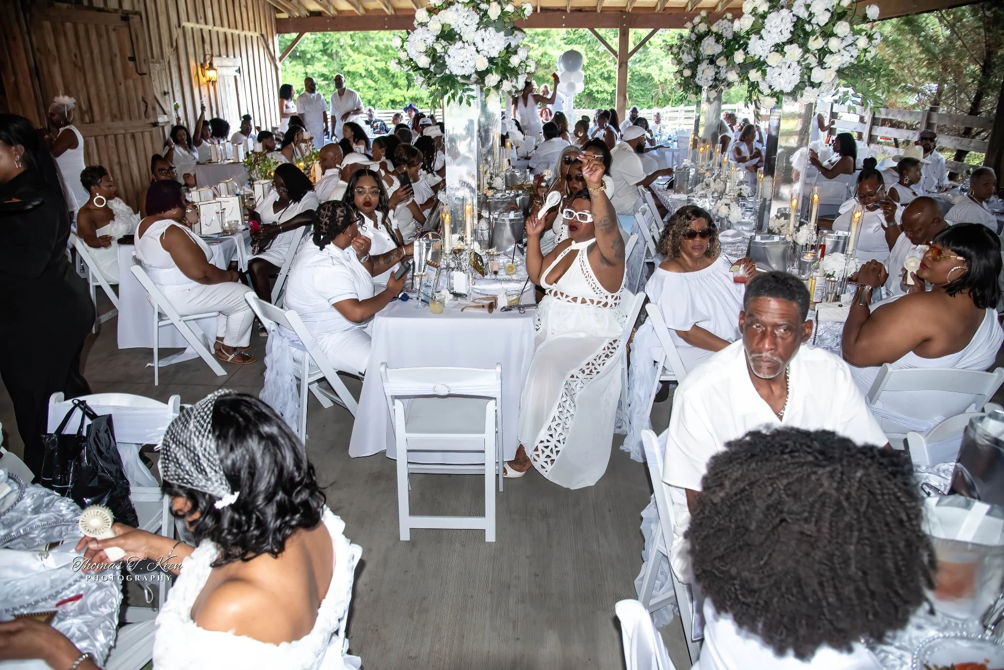 A large gathering of people dressed in white at a decorated outdoor event under a pavilion with floral arrangements and candles on the tables.