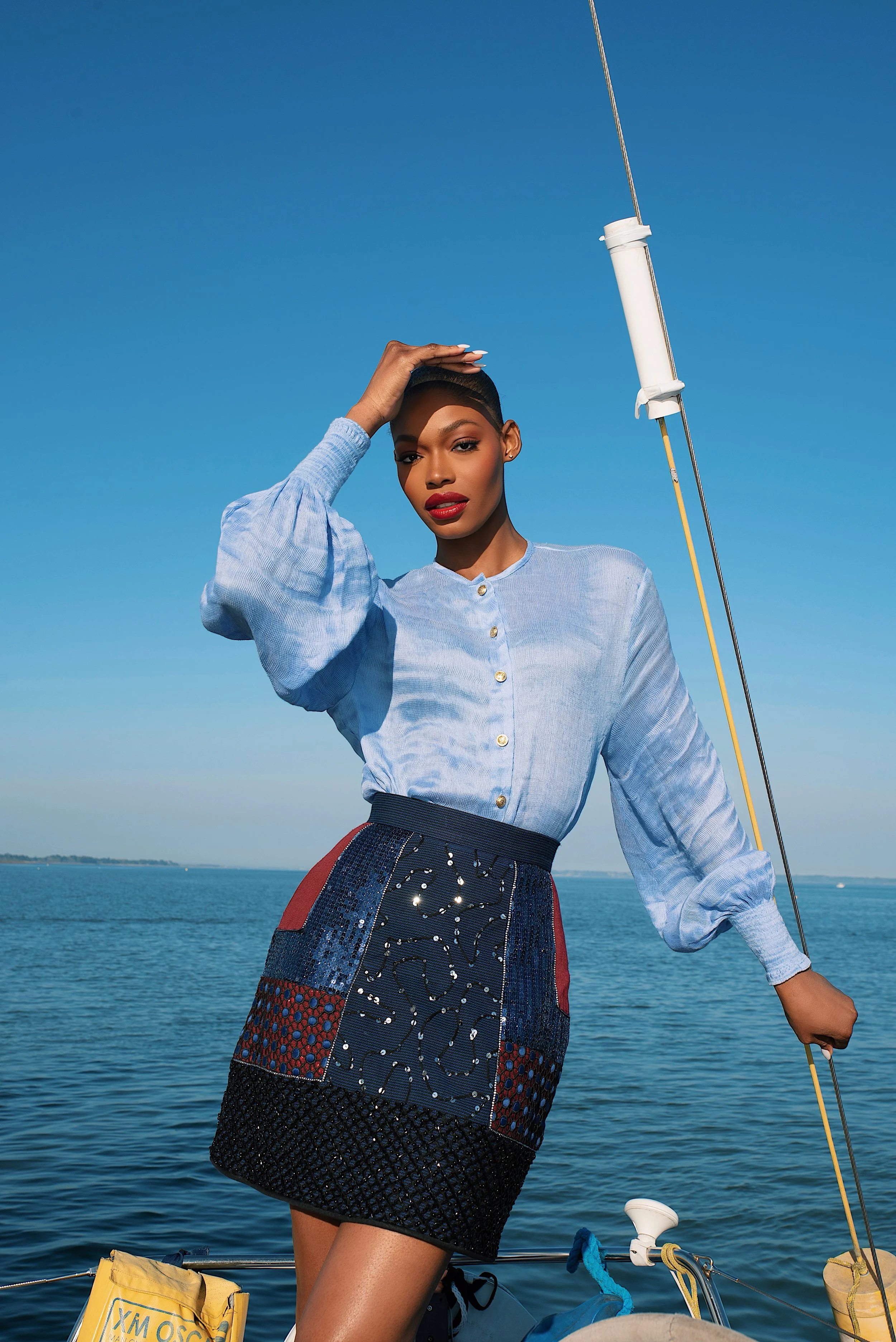 A woman in a light blue blouse and a patterned skirt stands on a boat by the water, holding a sailboat mast, with a clear blue sky in the background.