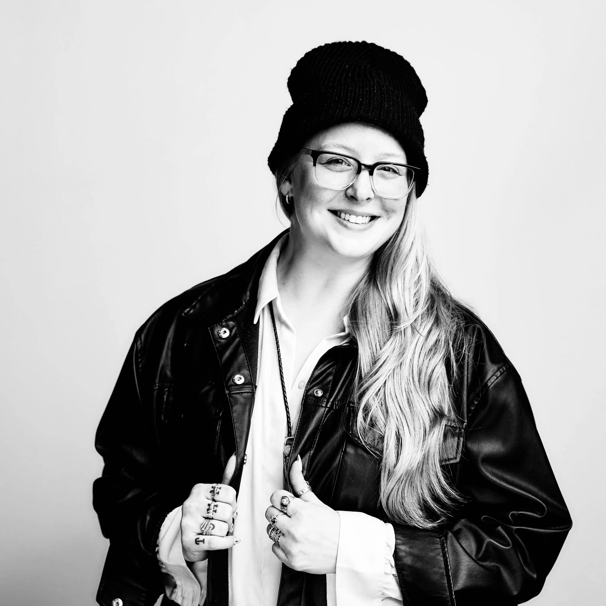 A woman with long wavy hair wearing glasses, a black beanie, a leather jacket, and a white shirt, smiling at the camera.