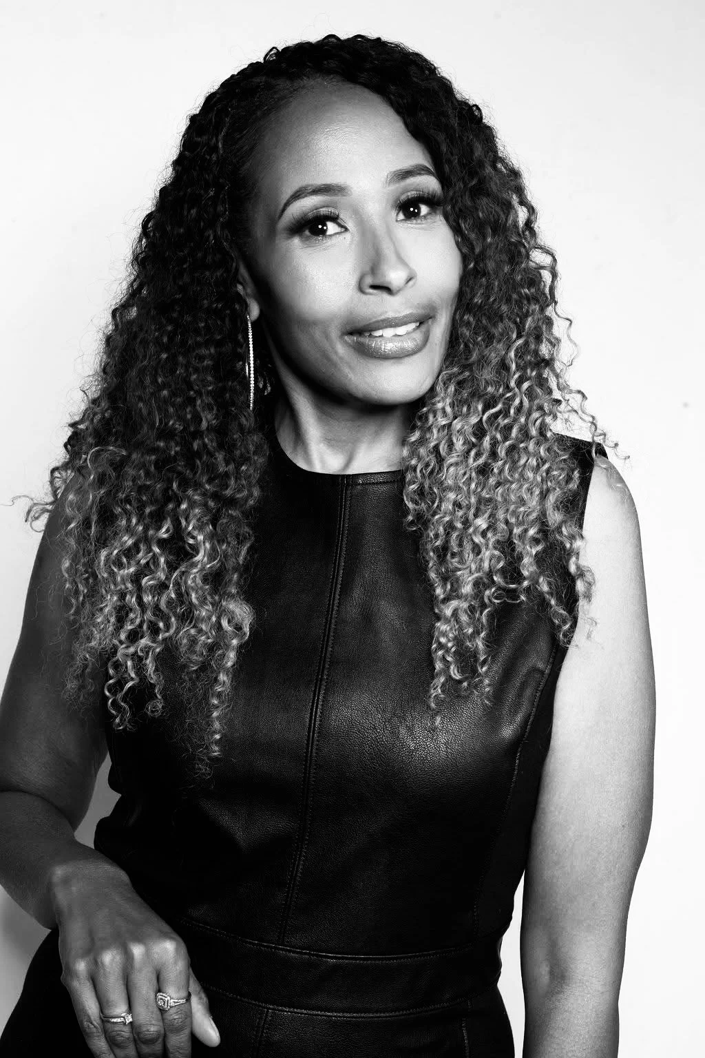 Black and white portrait of a woman with long, curly hair, wearing a sleeveless leather top and earrings.