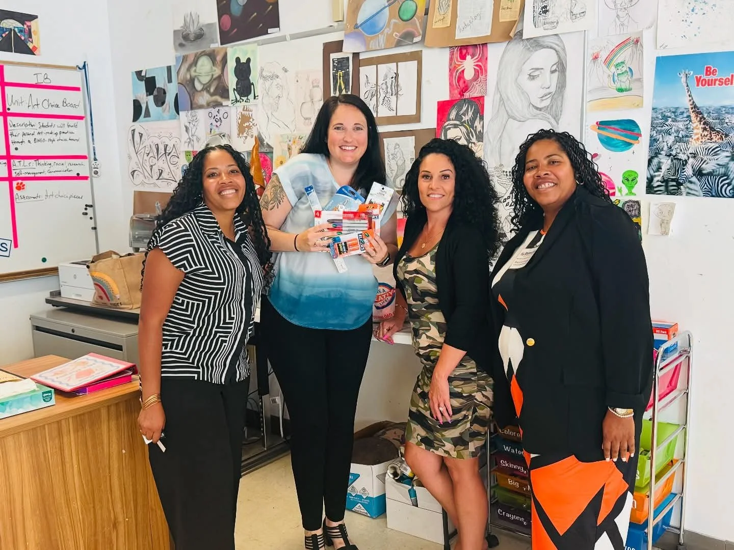 Four women standing together in a classroom, with art and drawings on the wall behind them. One woman is holding school supplies, including notebooks, folders, and sticky notes.