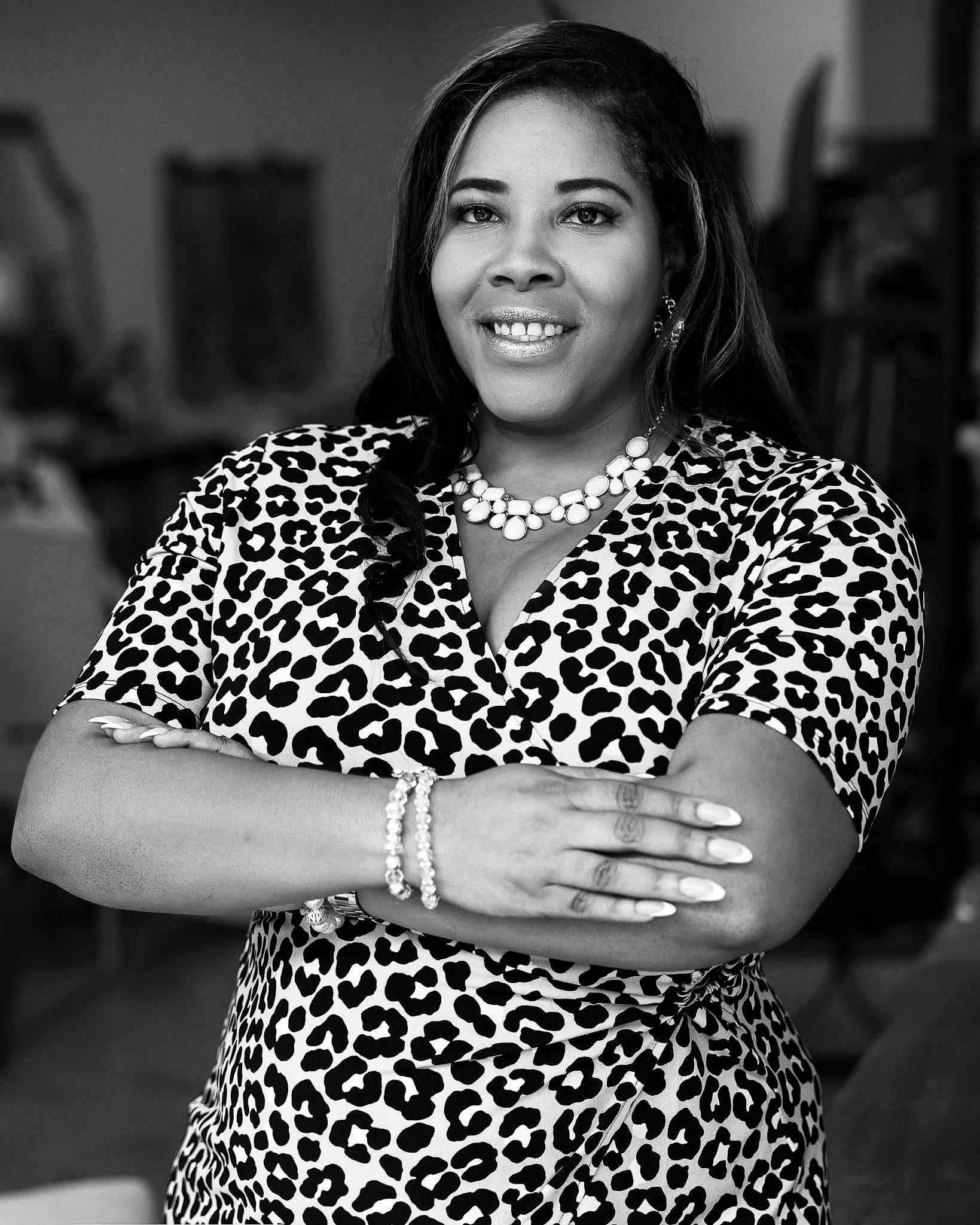 A portrait of a woman with medium skin tone, long dark hair, smiling, wearing a leopard print top, and jewelry including a pearl necklace and bracelet, standing with arms crossed.