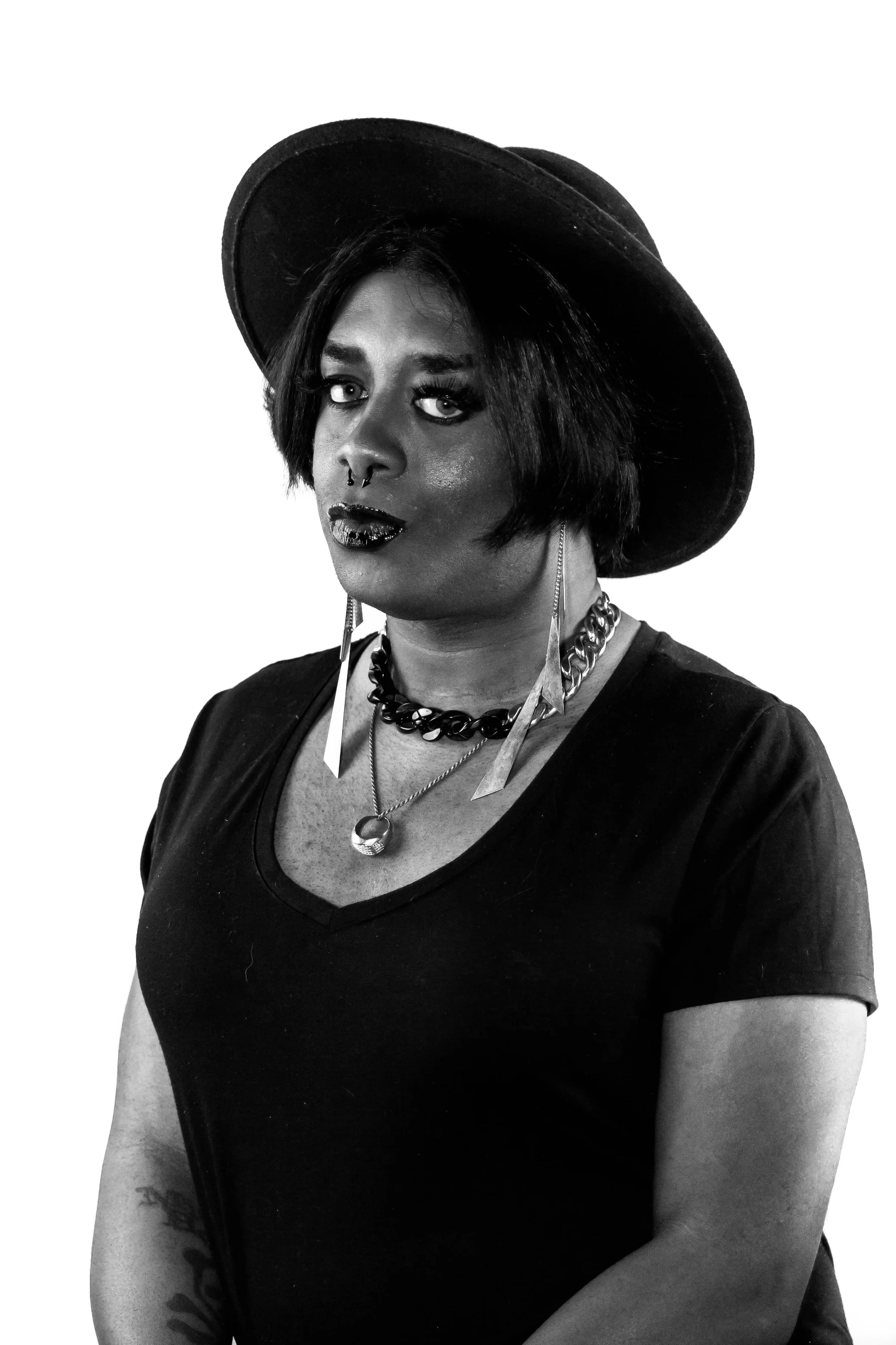 Portrait of a woman wearing a wide-brimmed hat, layered necklaces, and earrings, with dark makeup and a septum piercing, in black and white.