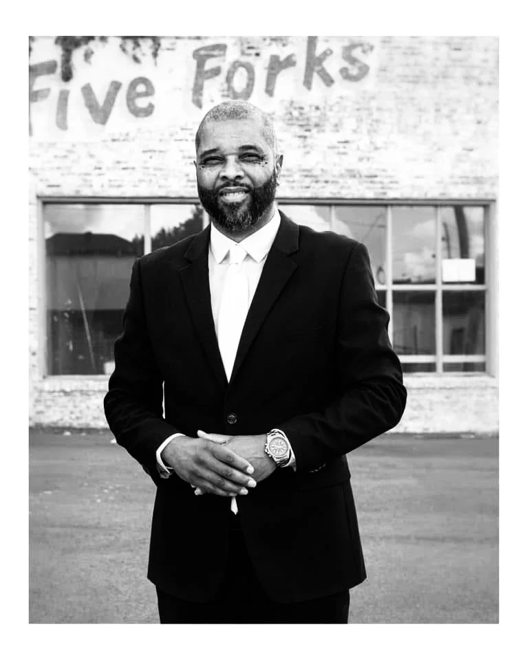 A man in a suit and tie with a beard, standing outdoors in front of a building with the words "Five Forks" partially visible in the background.