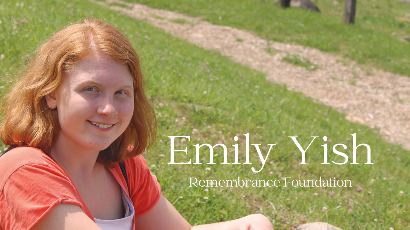 A young woman with red hair, wearing a red shirt, sitting outdoors on grass with a dirt path behind her. Overlaid text reads: Emily Yish, Remembrance Foundation.