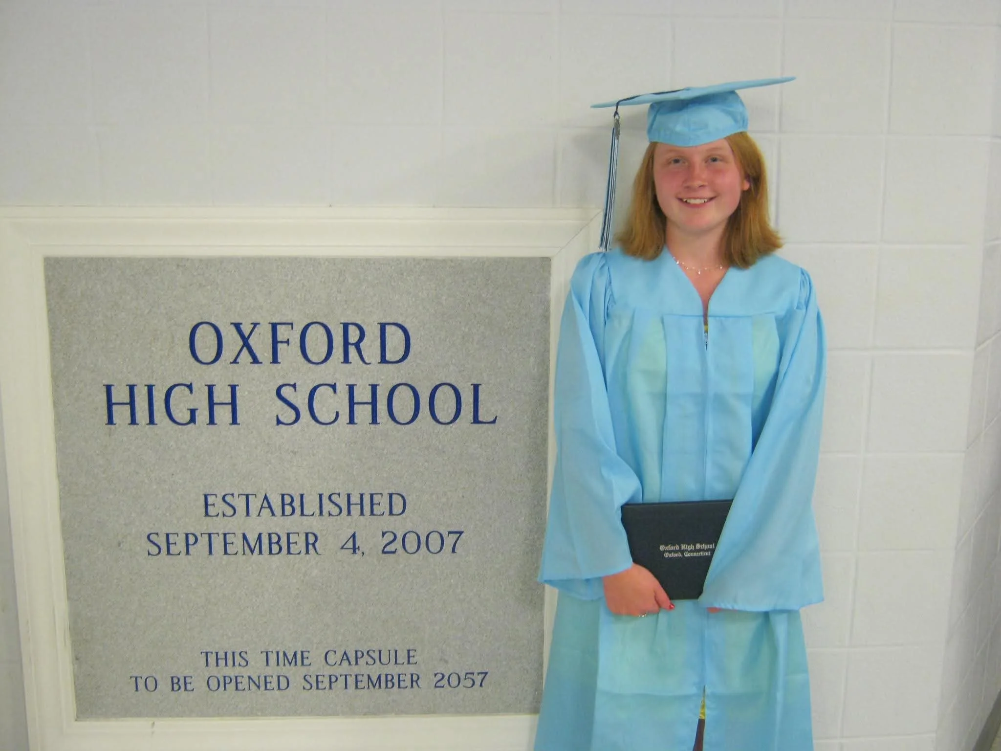 A young woman, Emily Yish,  in light blue graduation gown and cap holding a diploma, standing next to a sign that reads 'Oxford High School, established September 4, 2007, this time capsule to be opened September 2057'.