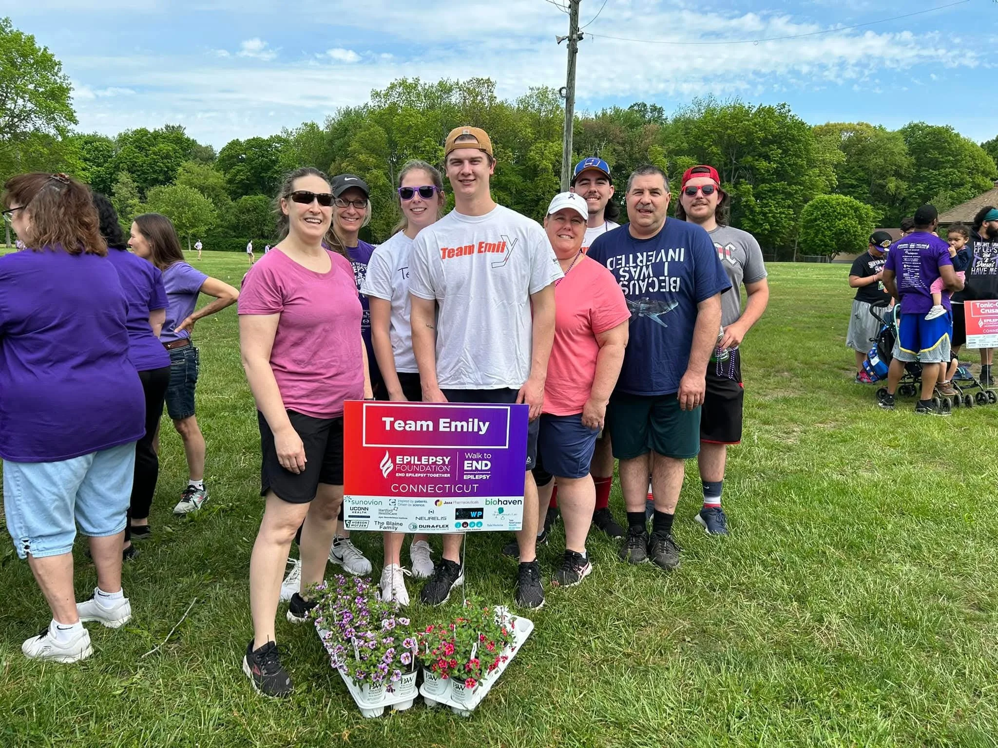 Group of people standing outdoors on grass, holding a sign that reads 'Team Emily' at a charity walk or event, with trees and a cloudy sky in the background.