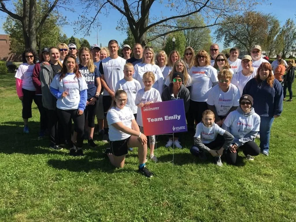 Group of people outdoors holding a sign that reads 'Team Emily' at a charity event for epilepsy awareness, with trees and a grassy field in the background.